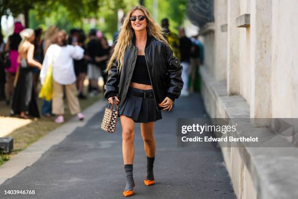 Emili Sindlev wears black sunglasses, a black shiny leather oversized bomber coat from Prada, a black shoulder-off cropped top, a dark gray pleated...