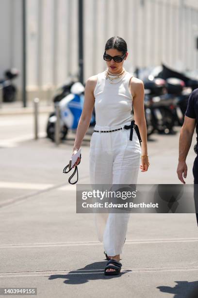 Caroline Issa wears black sunglasses, white pearls necklaces, a large gold chain necklace, a white ribbed halter-neck tank-top, a black chain belt...