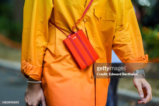 Pelayo Diaz wears black a tie and dye yellow to orange long blazer jacket, a red shiny leather crossbody bag from Sennei, a silver watch, a white...