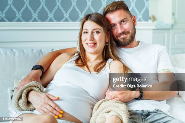 happy family of man and pregnant woman looking at camera with smile, hugging on a sofa at home - couple portrait soft stock pictures, royalty-free photos & images