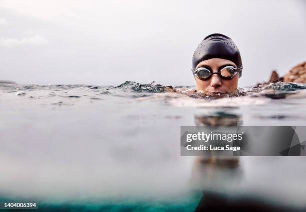 woman swimming towards camera, in the sea with swimming goggles and swim cap, body submerged. - simglasögon bildbanksfoton och bilder