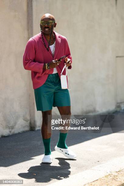 Guest wears dark green sunglasses, a white tank-top, a red ribbed pattern oversized shirt jacket, dark green suit shorts, a black Apple watch, gold...