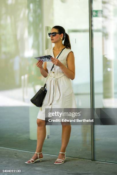 Guest wears black sunglasses, a white latte linen / sleeveless / belted short dress, a black shiny leather crossbody bag, white leather strappy...