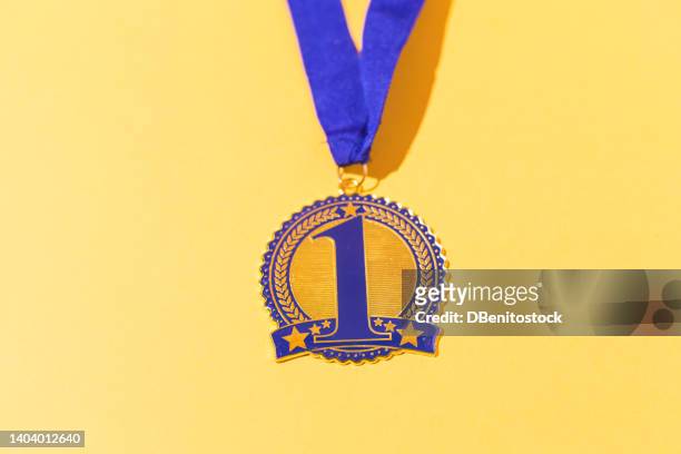 gold medal with blue decorations and the number 1 for sporting achievements for the first classified, on a yellow background. concept of winner, medals, honor and sports competition. - golden medals of merit in work ceremony stock pictures, royalty-free photos & images