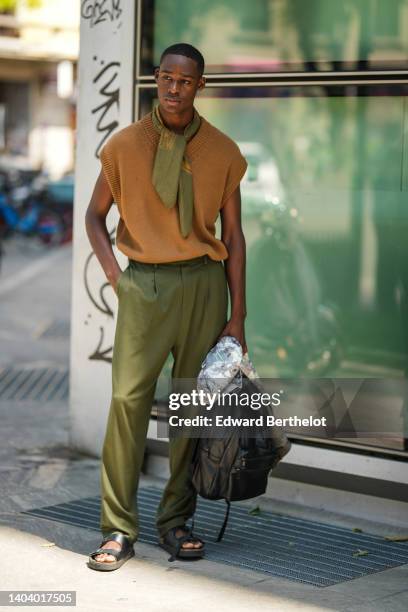 Guest wears a khaki and brown print pattern silk scarf, a brown wool sleeveless pullover, khaki large suit pants, a black shiny leather back pack...