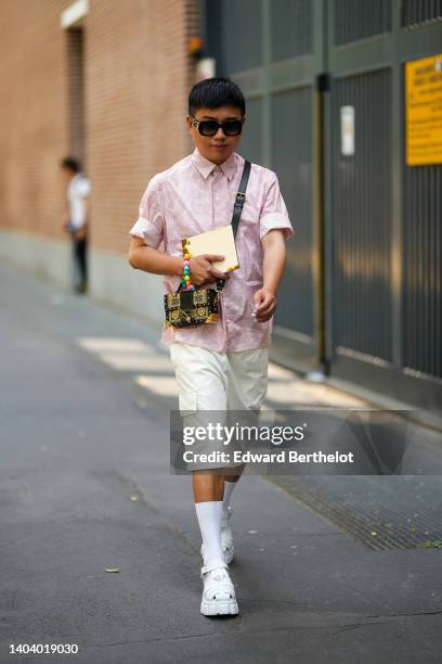 Guest wears black sunglasses, a pale pink and white print pattern short sleeves shirt, a black and gold print pattern crossbody bag, white cargo...