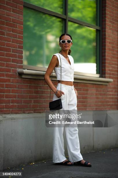 Guest wears white sunglasses, diamond earrings, a matching diamonds necklace, a white sleeveless cropped gilet, matching high waist flared suit...