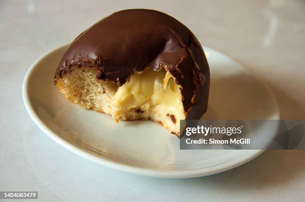 half-eaten chocolate iced vanilla bean cream puff on a round white plate on a cafe table - cream cake stock pictures, royalty-free photos & images