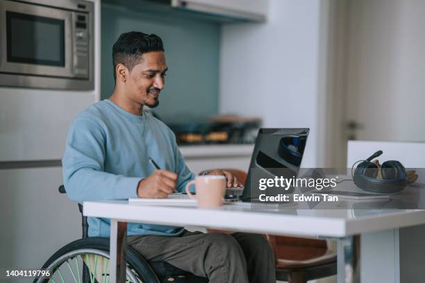 asian indian man in wheelchair sitting at dining room e-learning with laptop and writing notes - writer laptop stock pictures, royalty-free photos & images