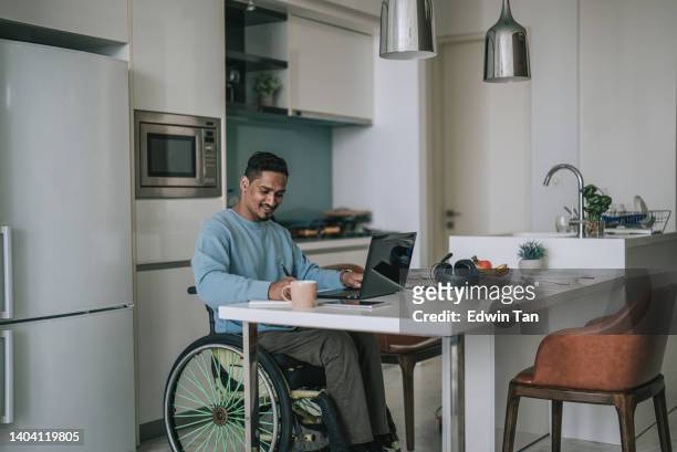 車椅子に乗ったアジア系インド人男性、食堂でノートpcでeラーニングをし、メモを書いている - mid adult ストックフォトと画像