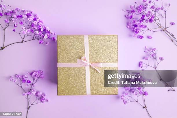 golden gift box with pink ribbon on purple background. purple flowers around box. - jubileum werk stockfoto's en -beelden