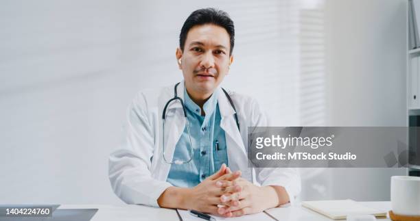 mature asian man doctors looking at camera video call medical results during consultation to patient in health clinic. - man talking to camera stock pictures, royalty-free photos & images