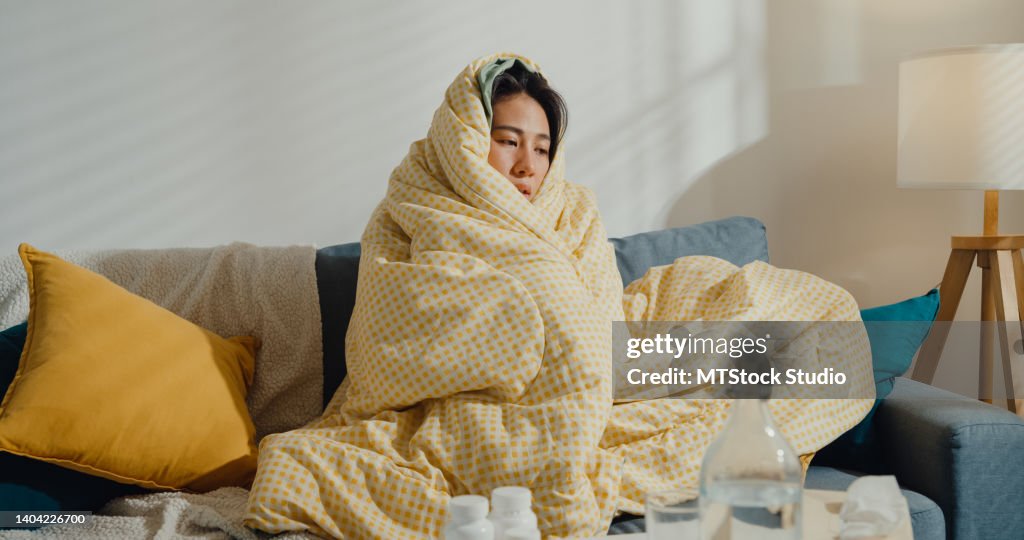 Kranke junge asiatische Frau Kopfschmerzen Fieber Husten Erkältung Sitzen unter der Decke auf dem Sofa im Wohnzimmer zu Hause.