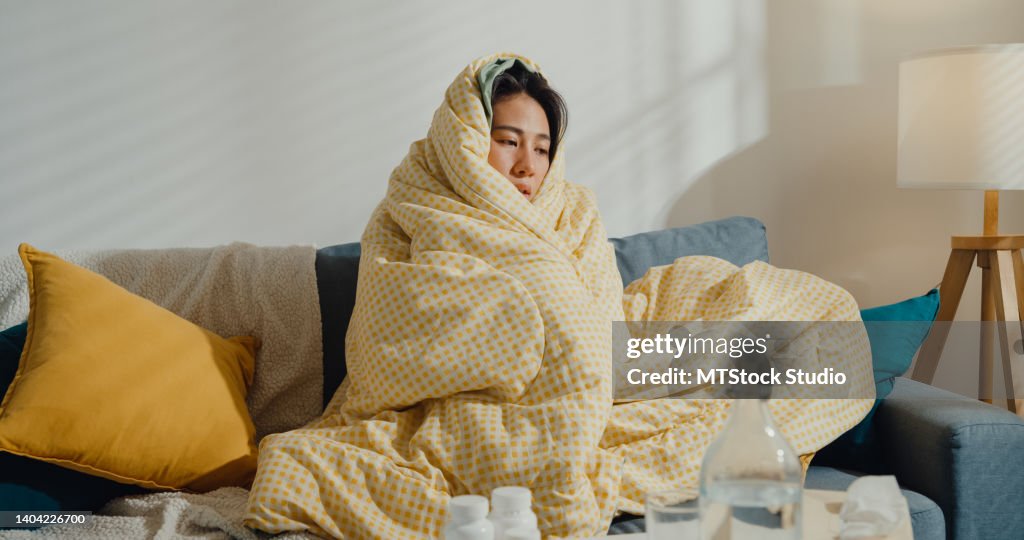 Sick young Asian woman headache fever cough cold sneezing sitting under the blanket on sofa in living room at home.