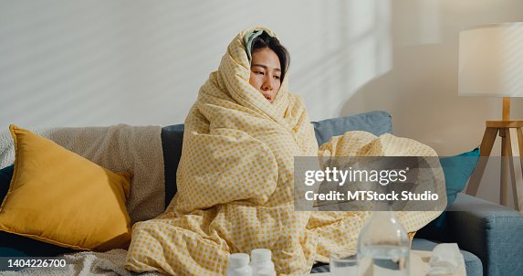 Sick young Asian woman headache fever cough cold sneezing sitting under the blanket on sofa in living room at home. Sick young Asian woman headache fever cough cold sneezing sitting under the blanket on sofa in living room at home.