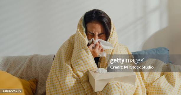 sick young asian woman blowing nose and sneeze with tissue paper sitting under the blanket on sofa in living room at home. - facial tissue stock pictures, royalty-free photos & images