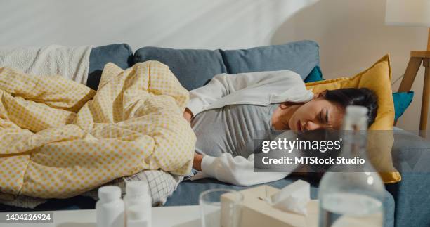 sick young asian woman suffering from strong abdominal pain lying down under the blanket on sofa in living room at home. - endometriosis stock pictures, royalty-free photos & images