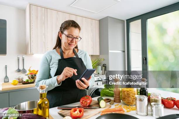 mujer cocinando y usando el teléfono móvil en la cocina - grabado objeto fabricado fotografías e imágenes de stock