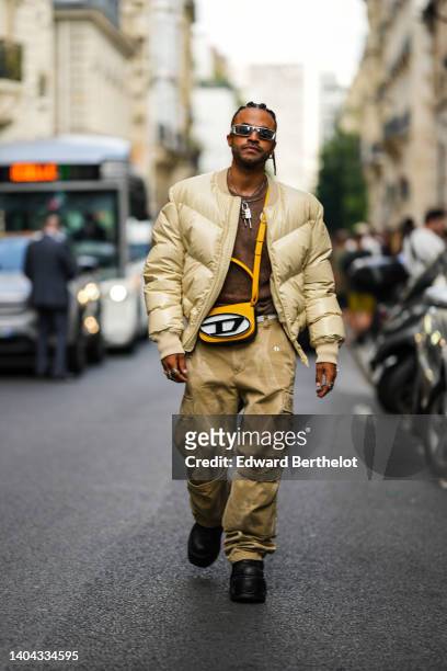 Theo Kimbaloula wears silver sunglasses, a brown t-shirt, a pale yellow oversized zipper puffer jacket, beige / pale yellow large cargo pants, a...