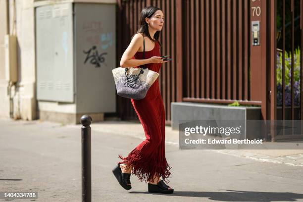 Clara Cornet wears gold and pearl pendant earrings, a gold large chain necklace, a burgundy ribbed / asymmetric shoulder / long fringed tube dress, a...