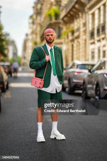 Karb wears a white shirt, a green silk tie, a dark green blazer jacket, matching dark green short suit, a pink shiny leather with white print pattern...