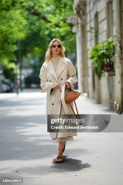 Natalia Verza wears black sunglasses from Prada, a beige linen long belted trench coat, a camel shiny leather with embossed pattern logo handbag from...