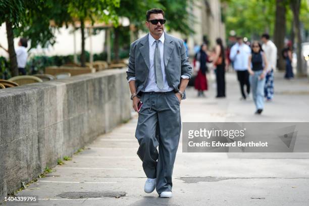 Alex Badia wears black sunglasses, a white shirt, a gray linen tie, a gray matching linen blazer jacket, gray matching suit pants, gold and silver...