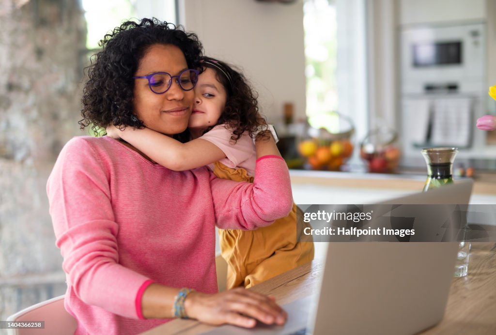 Multiracial girl with her mother hugging, having fun in kitchen during home-office and homeschooling time.