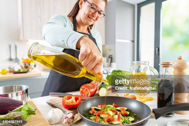 woman adding cooking oil to a frying pan with vegetables - olive oil stock pictures, royalty-free photos & images