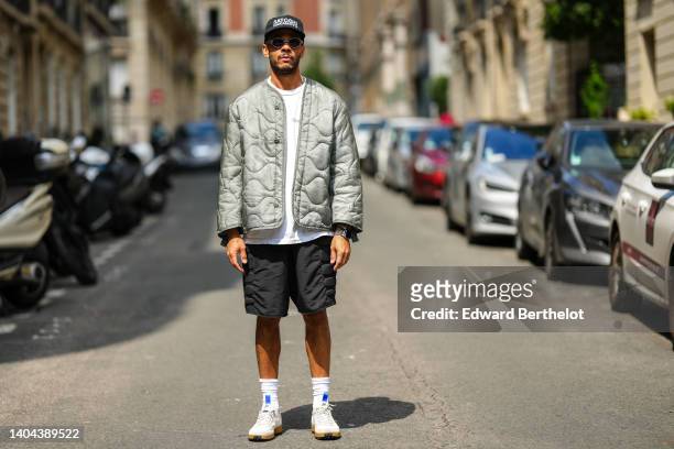 Guest wears a black cap with white slogan inscription "Satoshi Nakamoto", black circle sunglasses, white pearls necklaces, a white t-shirt, a silver...