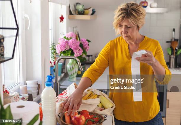 reife frau, die ihre quittungen zu hause durchgeht, nachdem sie lebensmittel gekauft hat - frau kauft lebensmittel stock-fotos und bilder