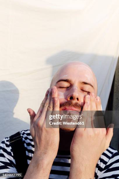 man with eyes closed applying moisturizer in front of white backdrop - hombre crema facial fotografías e imágenes de stock