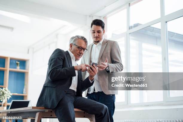 senior businessman sharing smart phone with young man in office - mid aged stock pictures, royalty-free photos & images