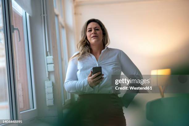 confident businesswoman with hand on hip holding smart phone at office - body types stock-fotos und bilder