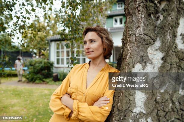 thoughtful woman with arms crossed leaning on tree trunk at back yard - person in front of house stock pictures, royalty-free photos & images