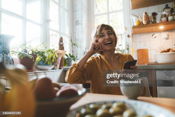 happy mature woman with smart phone sitting at dining table - woman kitchen stock-fotos und bilder