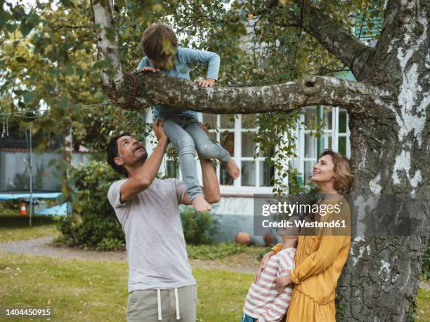 woman and girl looking at man helping boy climbing tree in back yard - kinder klettern stock-fotos und bilder