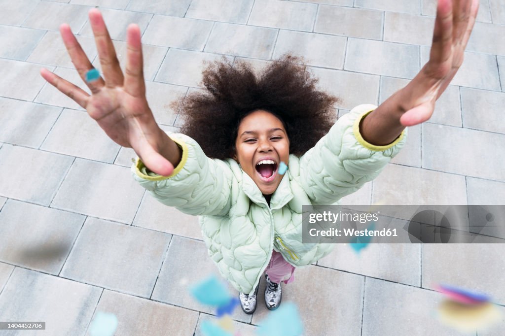 Cheerful girl with arms raised standing amidst confetti falling on footpath