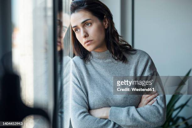depressed woman standing with arms crossed looking through window at home - 30 year old woman stock pictures, royalty-free photos & images