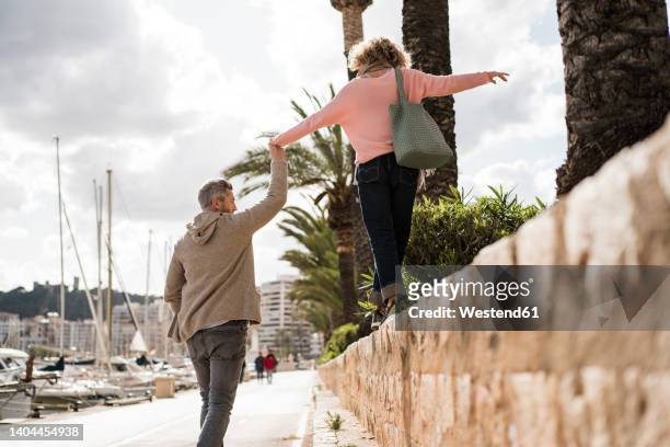 mature man holding hand of woman walking on wall by harbor - balancieren mauer stock-fotos und bilder