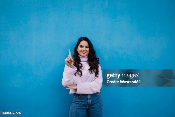 smiling young beautiful woman showing number one with index finger in front of blue wall - contare foto e immagini stock