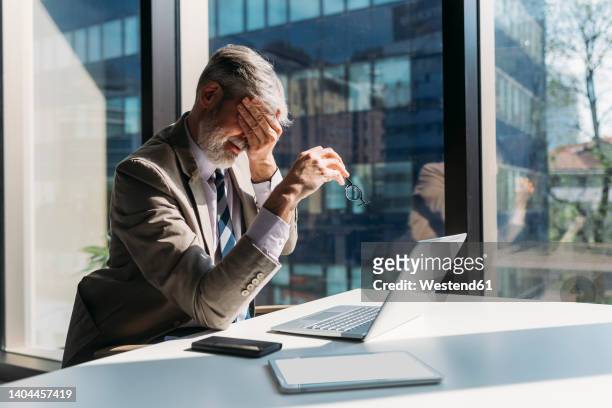 tired businessman sitting with hand covering eye at desk in office - augen zuhalten stock-fotos und bilder
