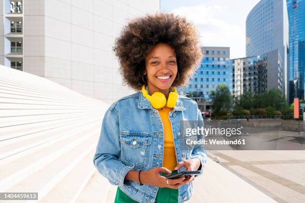 happy young woman wearing wireless headphones standing with smart phone by steps on sunny day - portrait of young woman standing against steps imagens e fotografias de stock