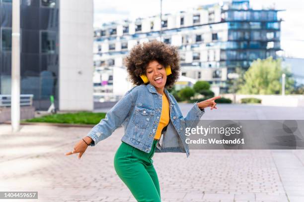 happy young woman listening music through wireless headphones enjoying dance on footpath - denim jacket fotografías e imágenes de stock