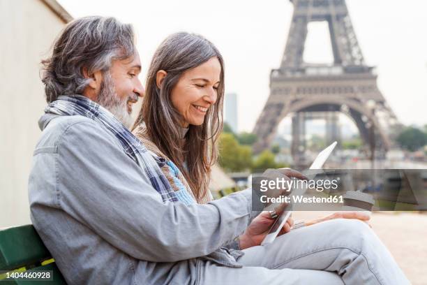 smiling mature woman with man using tablet pc sitting on bench near eiffel tower, paris, france - couple paris stock-fotos und bilder