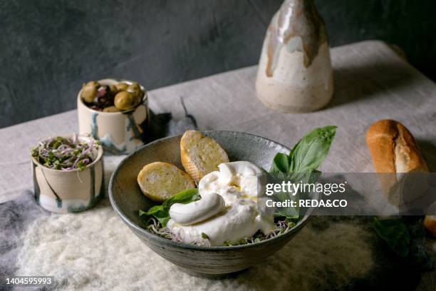 Traditional italian burrata knotted fresh cheese salad in grey ceramic bowl on table. Sliced bread, olives, green sprouts around. Healthy...