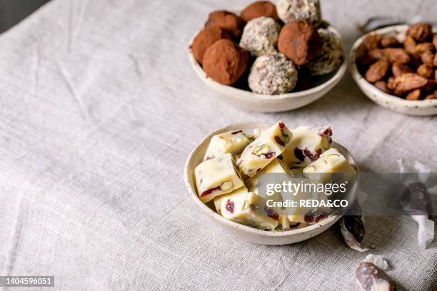 Collection of homemade sweet almond nuts, dark chocolate truffle, white chocolate candy bar in bowls on white linen table cloth. Handmade food sweet...