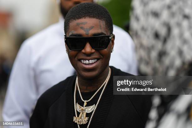 Kodak Black wears black sunglasses, diamond earrings, a black t-shirt, a black oversized cardigan, silver and gold bracelets, gold and diamonds...