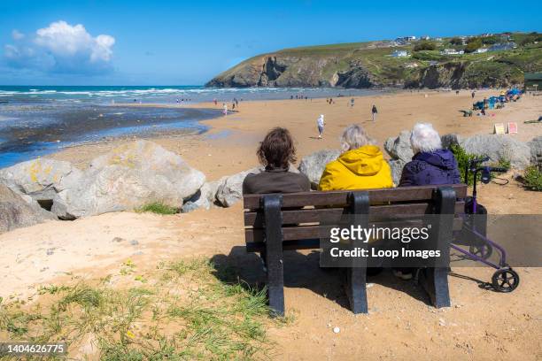 Mature holidaymakers sitting on a bench enjoying the sunny weather and the view over Mawgan Porth Beach in Cornwall.