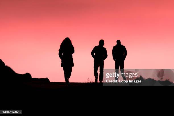 Three people walking along the coast path silhouetted against a colourful sky at the end of the day in Newquay in Cornwall.